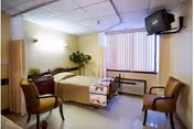 A nursing home bedroom with a single bed covered with a beige blanket and a patterned throw. There are two upholstered chairs, a wooden nightstand with a plant, a wall-mounted TV, a window with vertical blinds, and an air conditioning unit below the window.