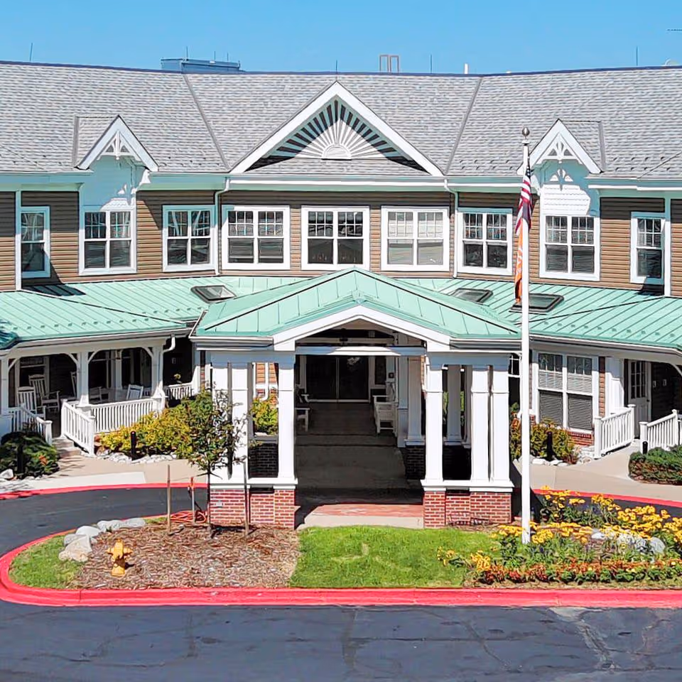 Front entrance of a multi-story senior living building with a covered porte-cochere, green metal roof, American flag, landscaping and a driveway.