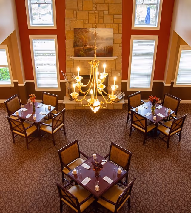 A cozy dining area with four square tables, each set with four chairs, napkins, and small vases with flowers. The room features a large stone fireplace with a painting above it, tall windows with blinds, and a decorative chandelier hanging from the ceiling.