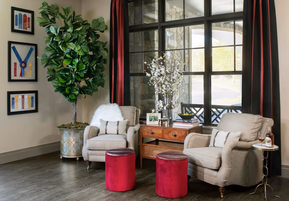 A cozy sitting area with two beige armchairs, each with a decorative pillow, positioned around a wooden side table with drawers. Two red velvet ottomans are in front of the chairs. A large potted plant is on the left side near framed ribbons on the wall. A window with dark red curtains shows an outdoor stone structure and greenery.