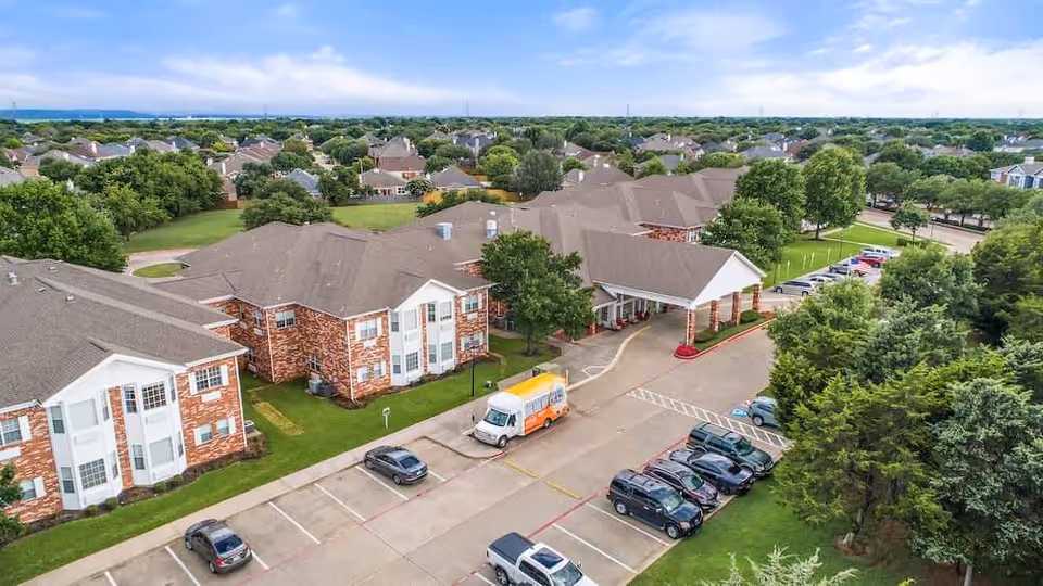 Aerial view of Morada Grand Prairie senior living facility showing multiple brick buildings with brown roofs surrounded by green trees and lawns. There is a parking lot with several cars and a shuttle van parked near the entrance of one building. The surrounding neighborhood with houses and trees is visible in the background under a cloudy sky.