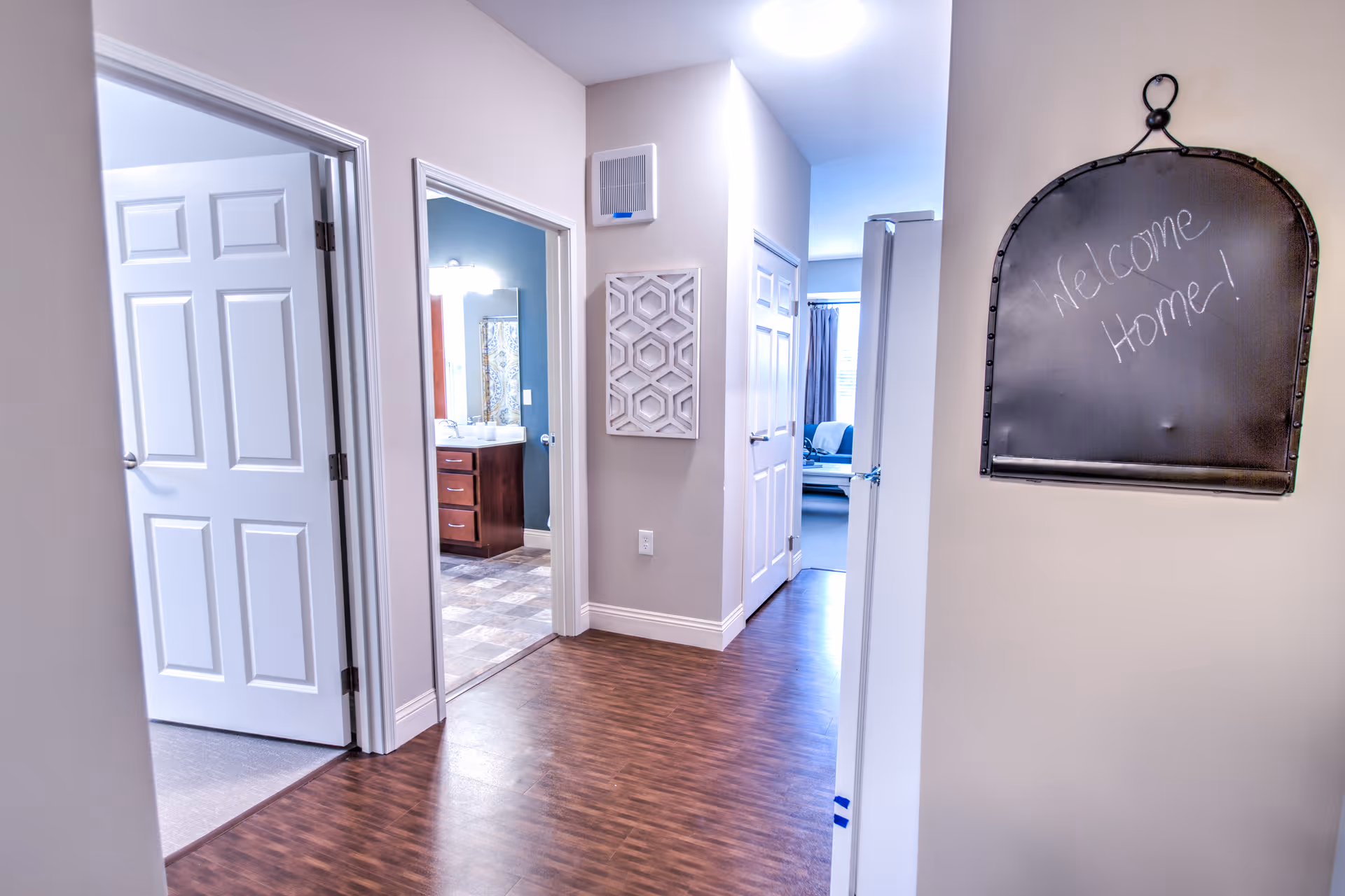 Interior hallway of a senior living facility with wooden flooring and light-colored walls. There are multiple open doors leading to different rooms, including a bathroom with a vanity and mirror visible. On the right wall, a blackboard with the handwritten message 'Welcome Home!' is hanging.