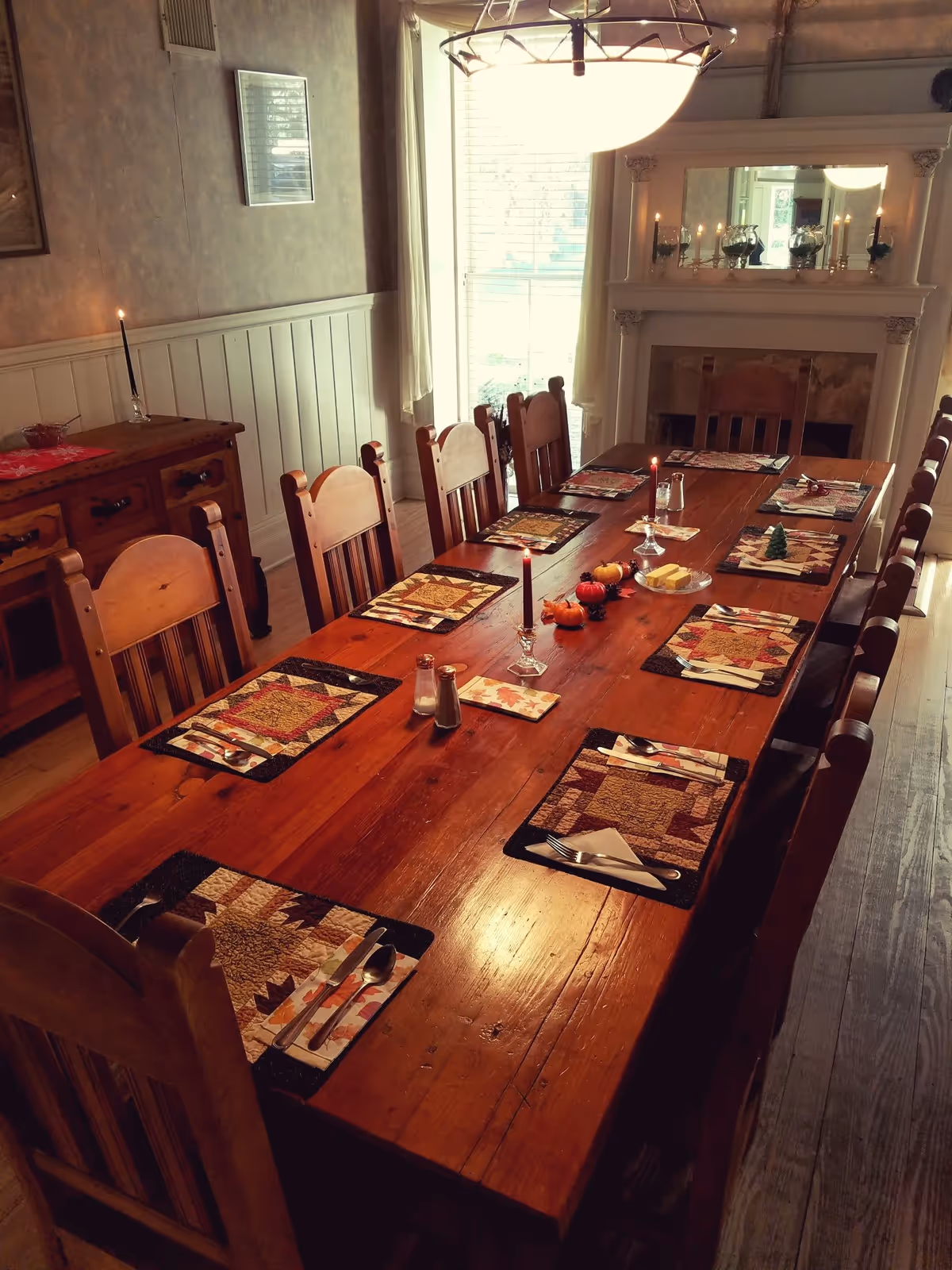A long wooden dining table set for eight with patterned placemats, silverware, and napkins. The table is decorated with small pumpkins, candles, and a plate of butter. The room features wooden chairs, a sideboard with a lit candle, a large window with curtains, and a fireplace with candles and decorative items on the mantel.