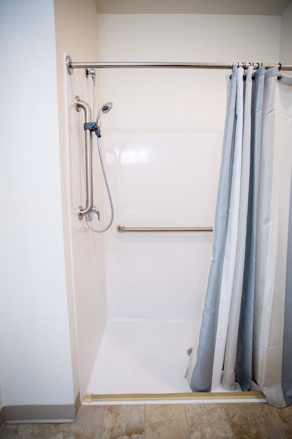 A clean, accessible shower stall with a handheld showerhead, grab bars on the wall, and a partially drawn light blue and white shower curtain. The floor outside the shower is tiled.