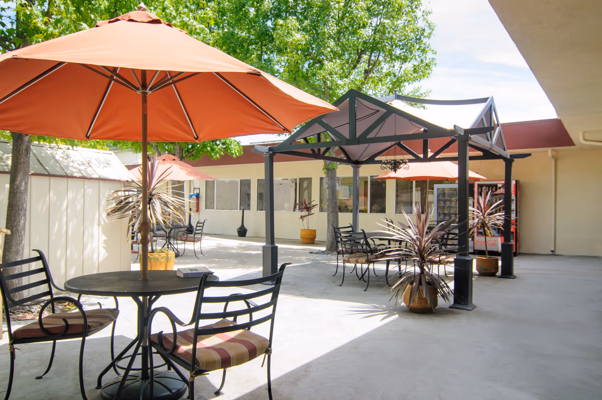 Outdoor courtyard with metal tables and chairs, orange umbrellas, potted plants and a pergola in front of a single-story building.