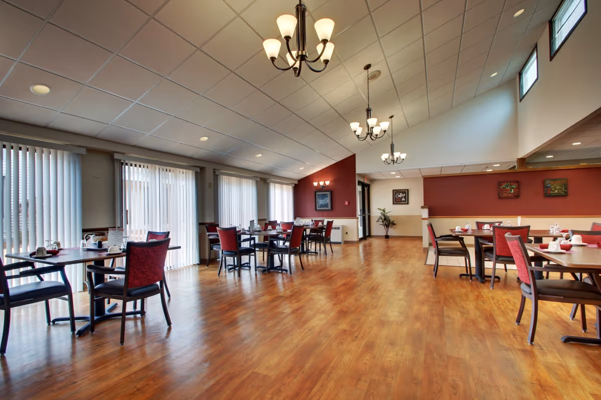 A spacious dining room with wooden floors, several tables and chairs with red upholstery, large windows with vertical blinds allowing natural light, and multiple ceiling light fixtures including chandeliers.