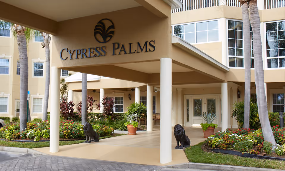 Entrance of Cypress Palms facility with a covered driveway supported by white columns, two lion statues on either side of the driveway, landscaped flower beds, palm trees, and a beige multi-story building in the background.