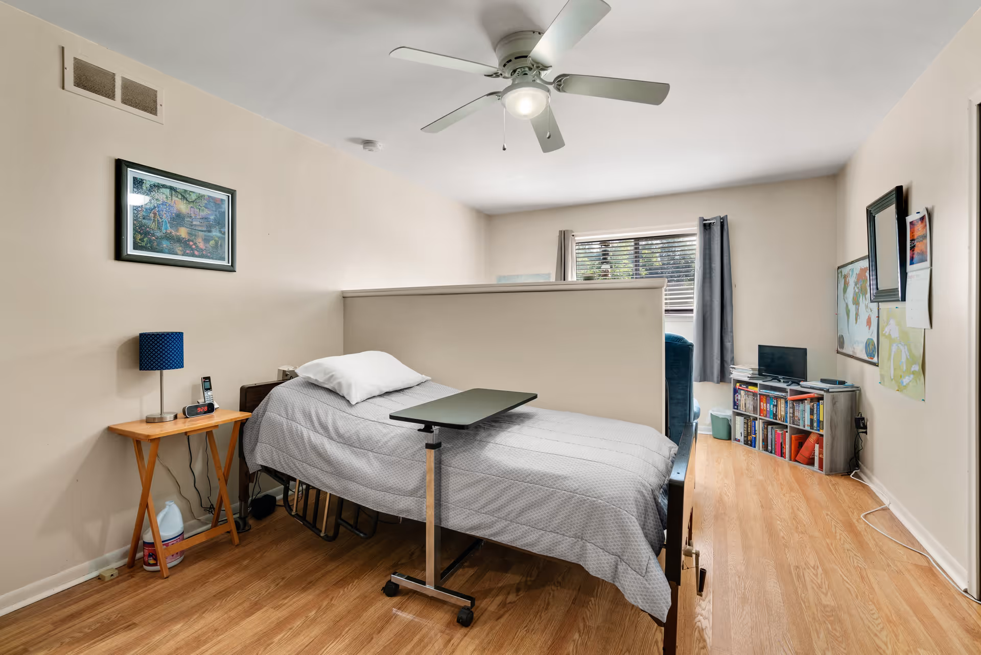 A simple bedroom with a single bed covered in a gray bedspread and a white pillow. Next to the bed is a small wooden side table with a blue lamp, a clock, and a phone. A small overbed table is positioned over the bed. The room has light-colored walls, a ceiling fan with a light, and a window with gray curtains. In the background, there is a small bookshelf with books and a TV on top, along with a map and a calendar on the wall.