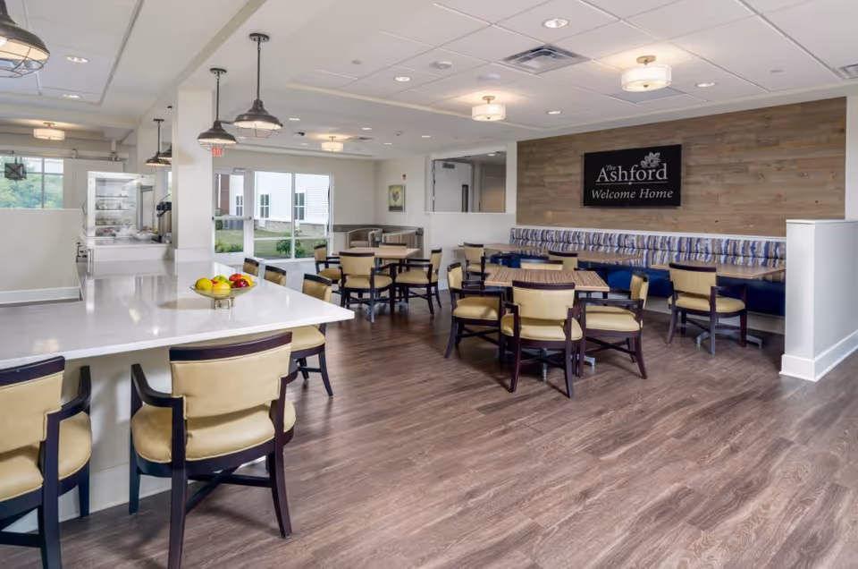 A bright and spacious dining area in The Ashford of Grove City featuring multiple tables and chairs arranged on a wood floor. There is a long counter with chairs on one side and a bowl of fruit on the counter. The back wall has a wooden panel with a sign that reads 'The Ashford Welcome Home'. Large windows allow natural light to fill the room.