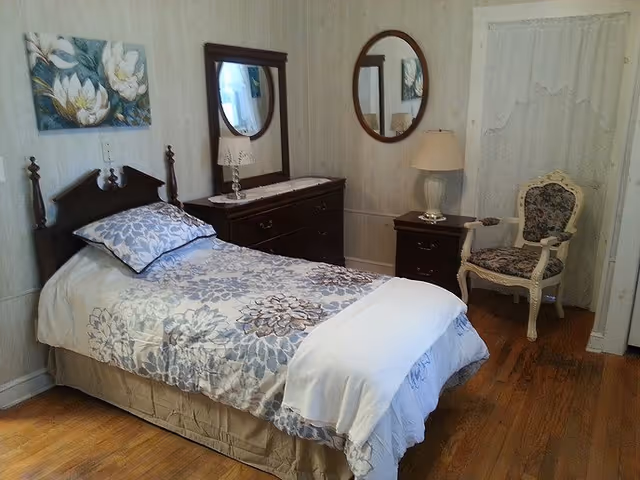 A cozy bedroom with a single bed covered in a floral patterned bedspread and pillow. There is a wooden dresser with a mirror above it, a bedside table with a lamp, and an upholstered armchair with a floral design. The room has wooden flooring and light-colored walls with a round mirror hanging on one wall.