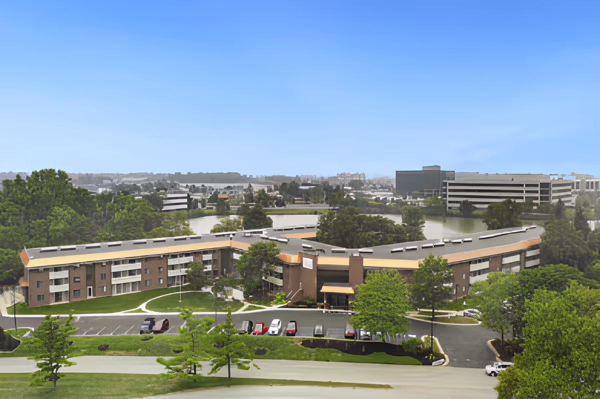 Aerial view of a curved three-story senior living building surrounded by lawns, trees, parking and a lake with office buildings in the distance.