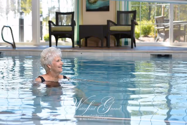 An elderly woman with short white hair is smiling and standing in an indoor swimming pool. Behind her, there are two chairs with cushions and a small table between them. Large windows let in natural light and show an outdoor view with trees and patio furniture.