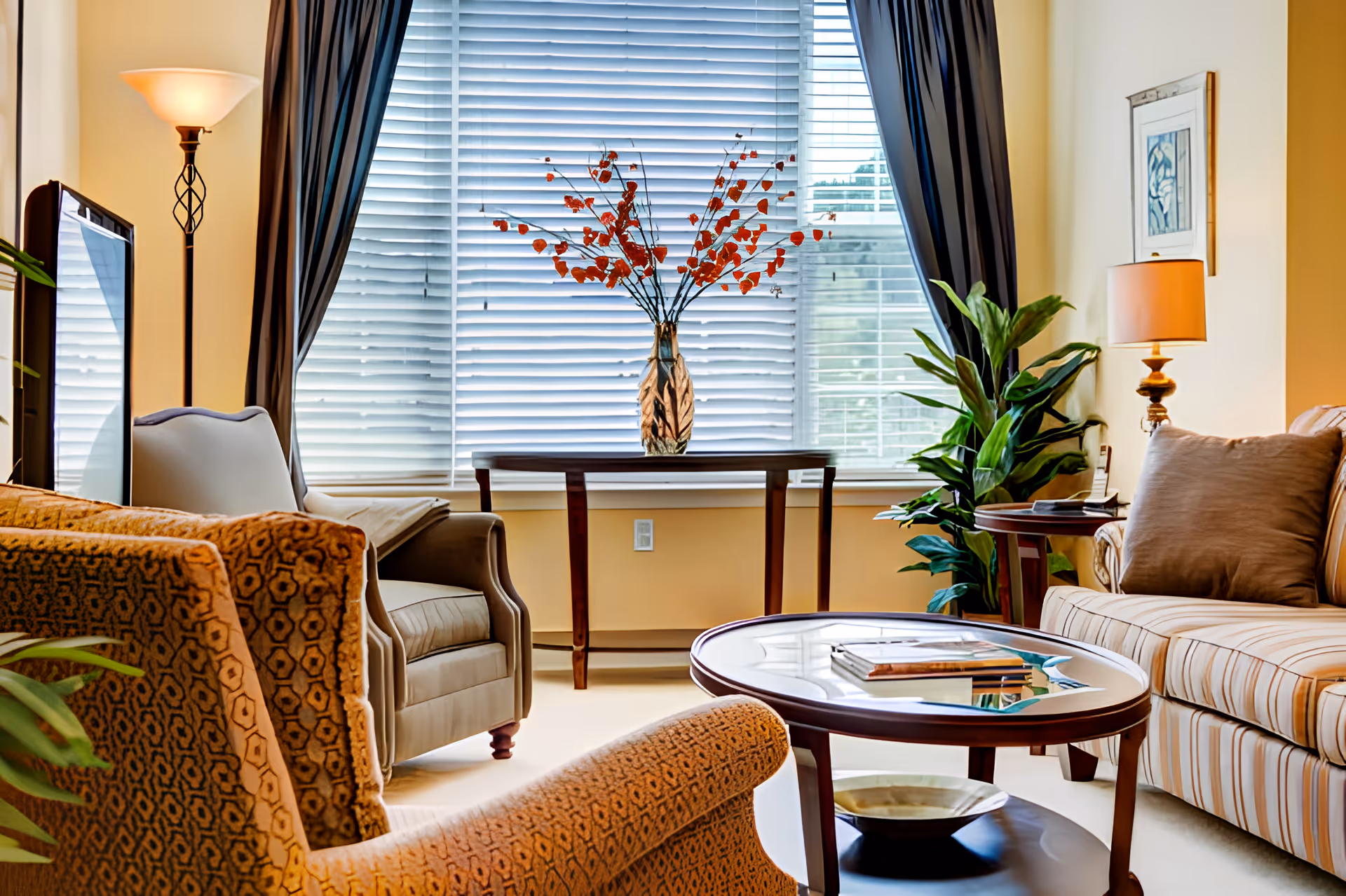 A cozy living room with a patterned armchair, a beige armchair, and a striped sofa with cushions. A round glass coffee table with books and a decorative bowl sits in the center. Behind, a wooden console table holds a vase with orange flowers in front of a large window with closed blinds and dark curtains. There are two lamps, one floor lamp and one table lamp, and a green potted plant near the sofa.