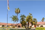 Exterior view of a single-story building with a red-tiled roof, surrounded by palm trees and a well-maintained lawn under a clear blue sky. An American flag and another flag are visible on a flagpole in front of the building.