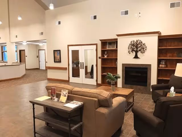 Seating area with a sofa and armchairs arranged around a coffee table facing a fireplace and built-in shelving in a spacious common room.