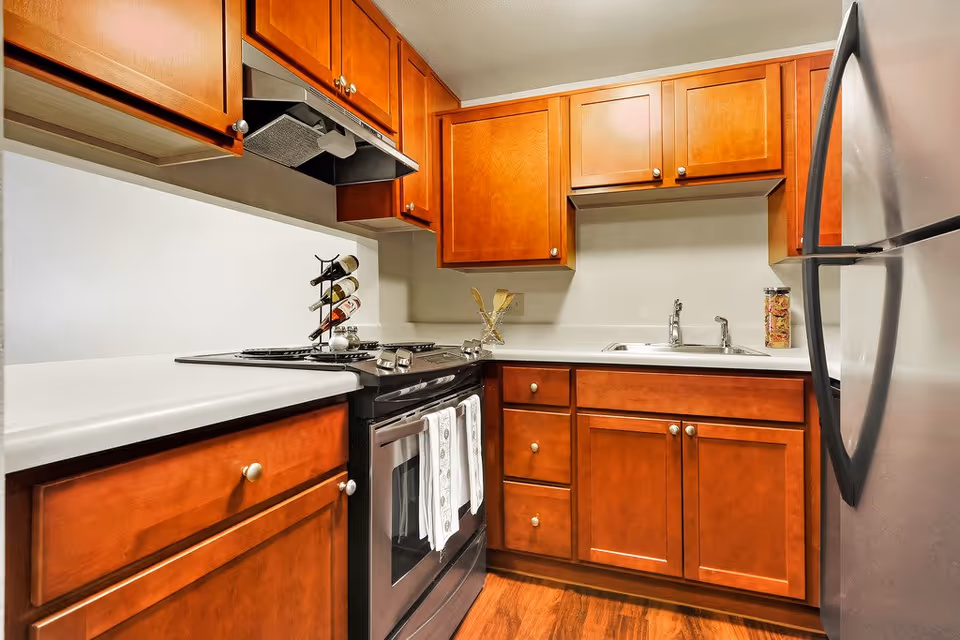 A compact kitchen with wooden cabinets, a black stove with two white towels hanging on the handle, a stainless steel refrigerator, a double sink, and a wine rack holding three bottles on the countertop.
