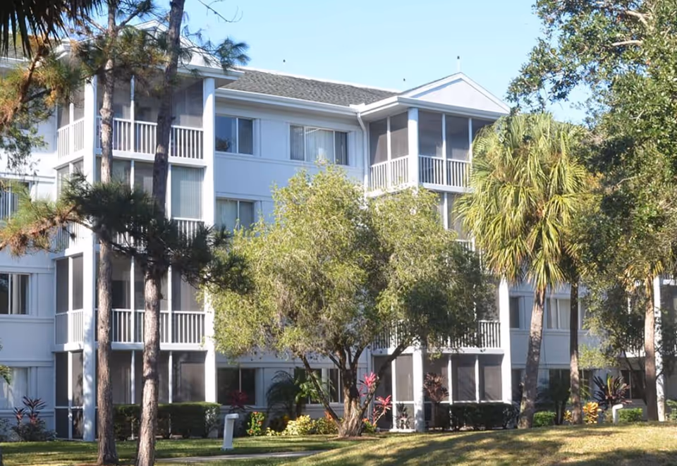 Exterior view of a multi-story assisted living facility building surrounded by trees and greenery under a clear blue sky.