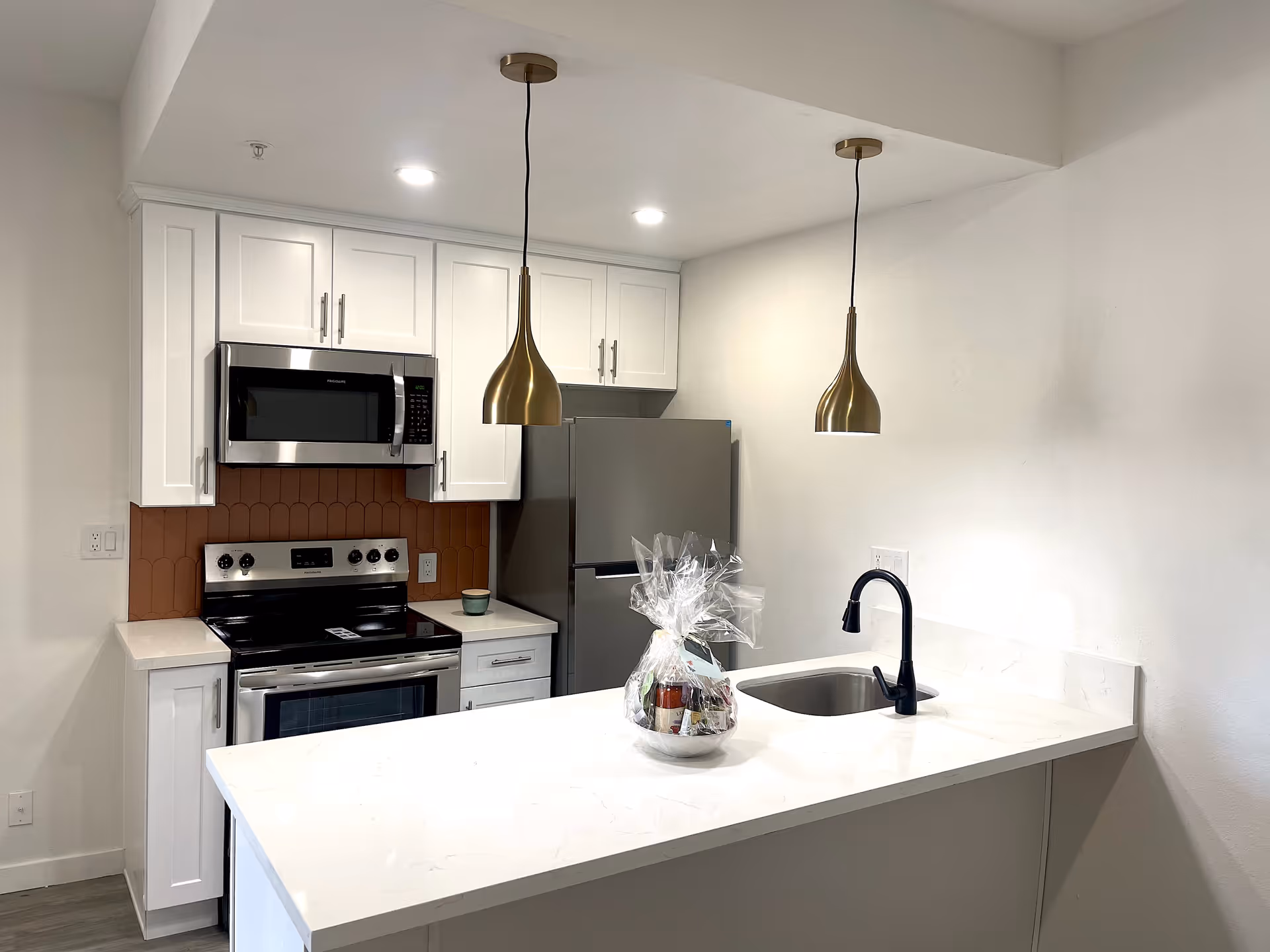 Modern kitchen with white cabinets, stainless steel appliances including a microwave, stove, and refrigerator. There is a white countertop island with a black faucet and sink, two gold pendant lights hanging above, and a small gift basket wrapped in clear plastic on the counter.