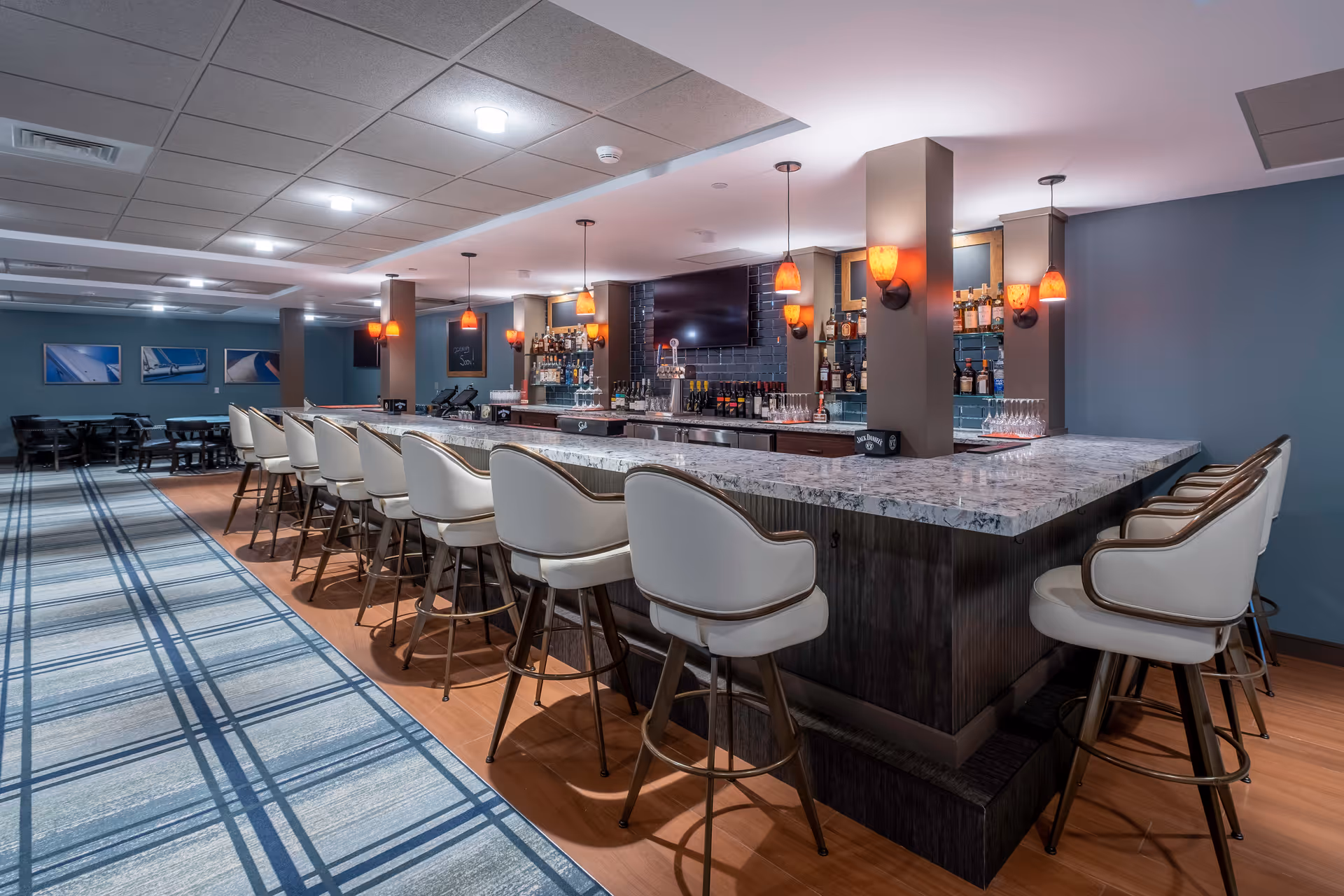 Interior view of a modern bar area with a long marble countertop surrounded by white cushioned bar stools. The bar is well-lit with hanging orange pendant lights and wall sconces. Behind the bar, shelves are stocked with various bottles and glassware. The floor has a combination of wood and a blue patterned carpet, and there are tables and chairs in the background along the wall with framed pictures.