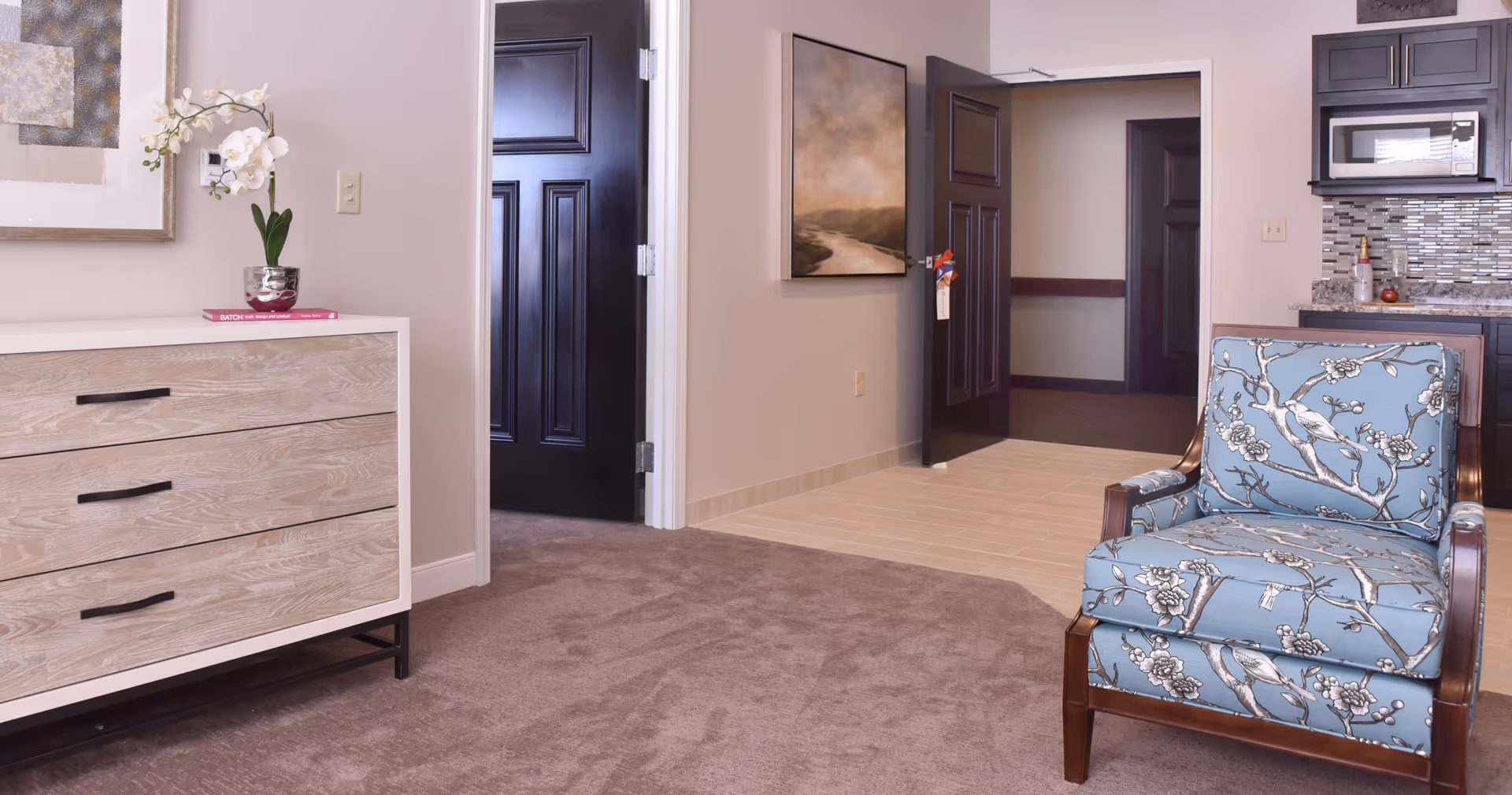 Interior view of a senior living facility room at Wooded Glen showing a patterned blue armchair, a wooden dresser with a small plant and books on top, two black doors, a painting on the wall, and a kitchenette area with a microwave and tiled backsplash.