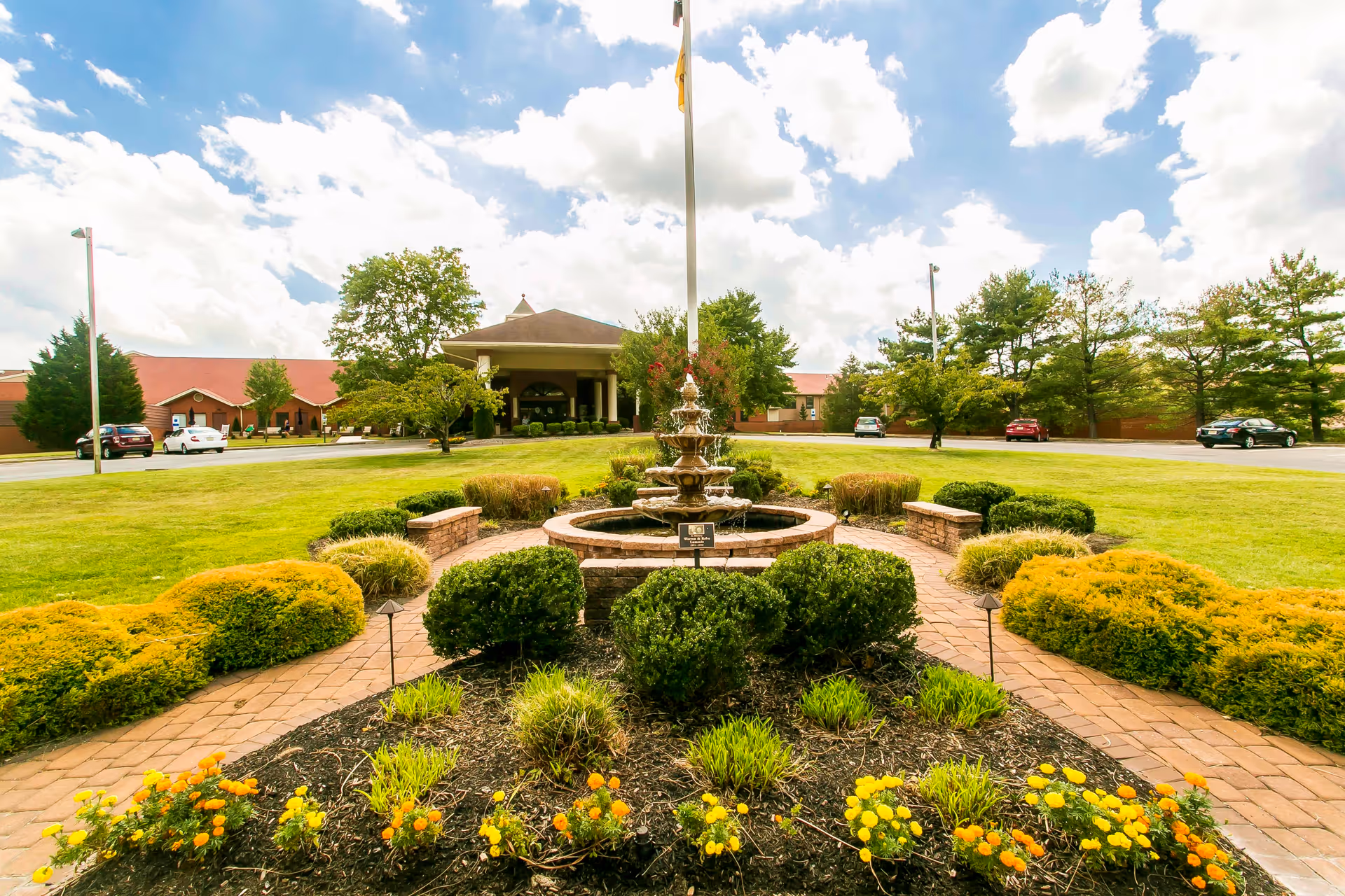 A landscaped garden area with a central multi-tiered water fountain surrounded by neatly trimmed bushes and yellow flowers. A brick pathway leads to the fountain, and in the background is a large building with a covered entrance and a red roof under a partly cloudy sky.
