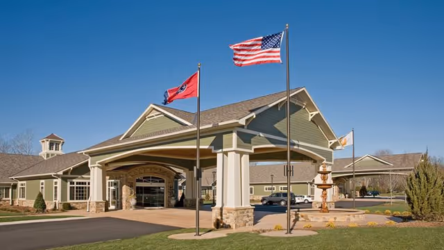 Exterior view of a senior living facility with a covered entrance, two flagpoles displaying the American flag and a state flag, a fountain, and a clear blue sky.