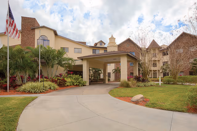 Entrance to a senior living facility with a covered drop-off, landscaped lawn, and a flag.