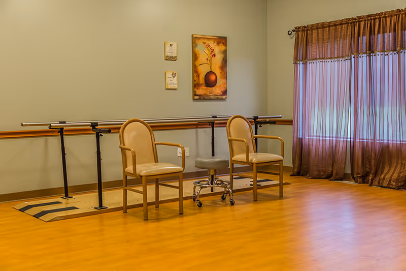 A rehabilitation room with two beige chairs and a gray rolling stool placed in front of parallel bars used for physical therapy. The room has wooden flooring, a window with brown curtains, and three pieces of wall art including a painting of a vase with flowers.