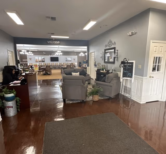 Lobby-style common area with a reception desk on the left, a seating group of armchairs in the center, and a dining/activity area visible beyond a 'welcome' sign.
