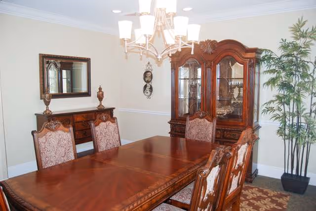 A traditional dining room with a polished wooden dining table and six upholstered chairs. The room features a wooden china cabinet with glass doors displaying decorative items, a sideboard with a mirror above it, a chandelier with multiple lights hanging from the ceiling, and a tall potted plant in the corner.