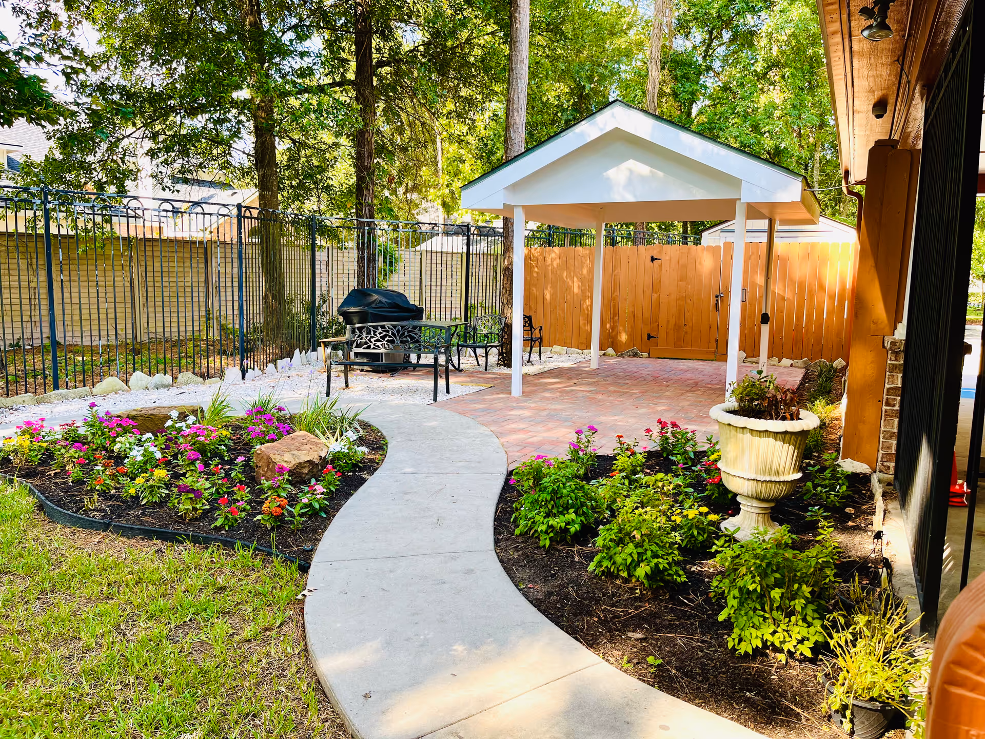 Curved concrete path winds through a landscaped courtyard with colorful flower beds, a covered patio, seating and a grill.
