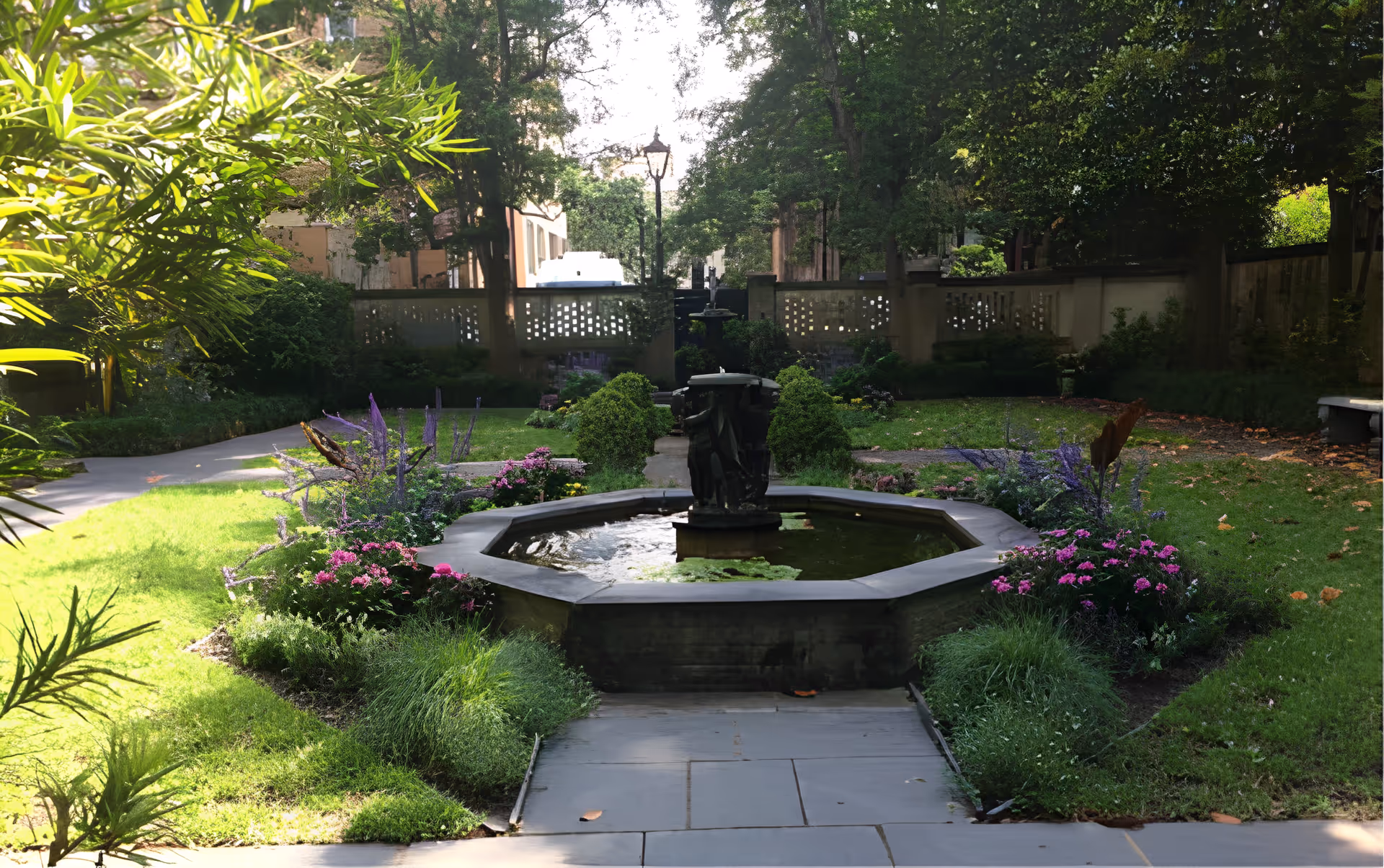 A peaceful outdoor garden area with a central octagonal water fountain surrounded by green grass, flowering plants, and trees. There is a paved walkway leading to the fountain, and a stone bench is visible on the right side. The garden is enclosed by a decorative wall and shaded by tall trees.