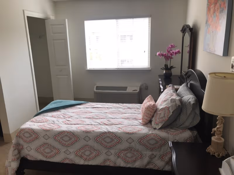 A neatly made bedroom with a patterned bed, nightstand and lamp, a dresser with flowers, and a window above an air-conditioning unit.