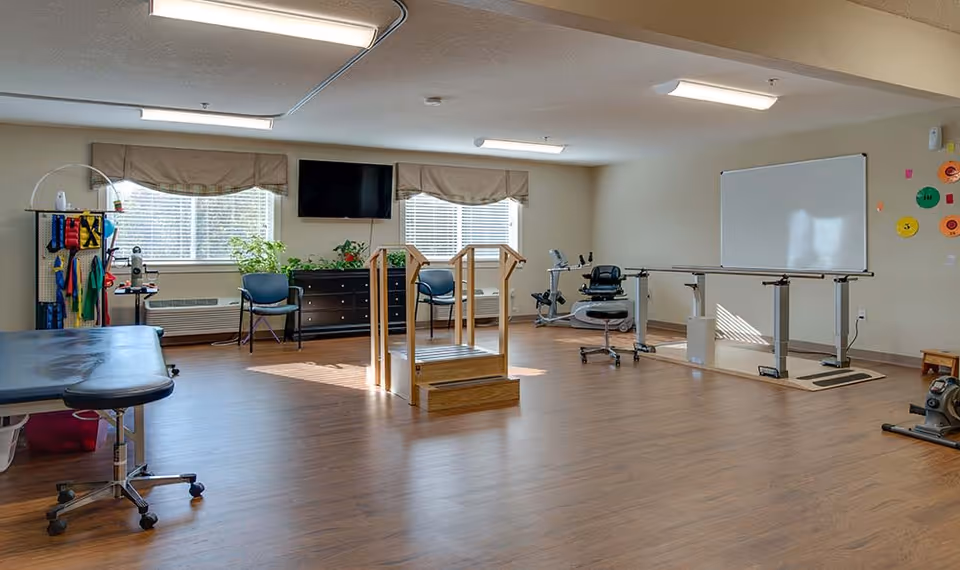 Spacious interior therapy/exercise room with parallel bars, treatment table, exercise equipment, TV and seating.