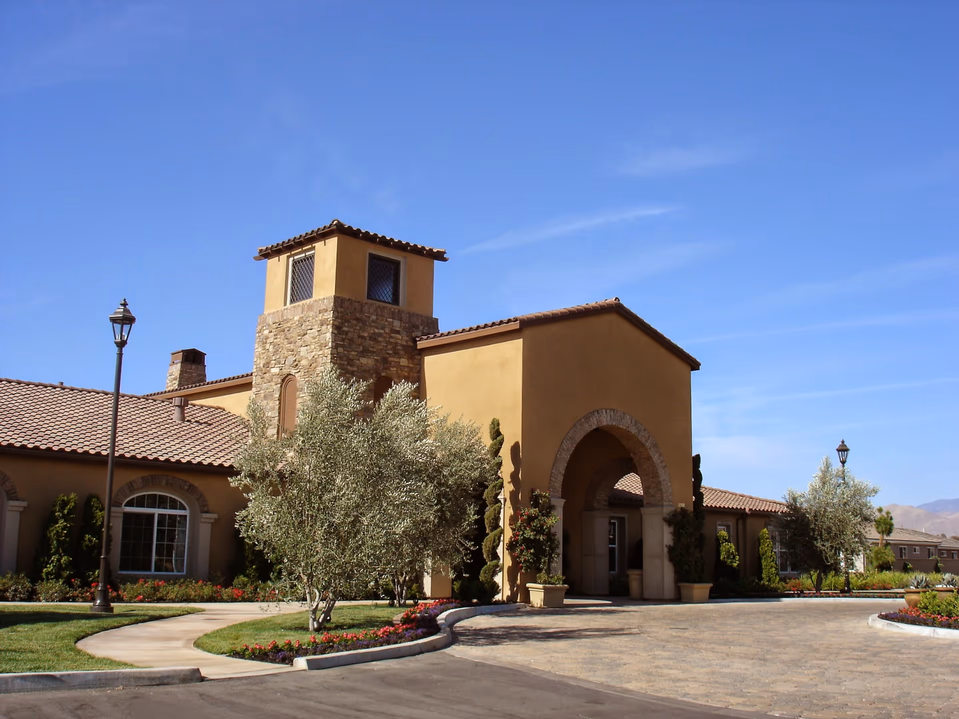 Exterior view of Four Seasons at Bakersfield Recreation Center featuring a Mediterranean-style building with tan stucco walls, a stone tower, arched entrance, tiled roof, landscaped garden with trees and flowers, and a clear blue sky.