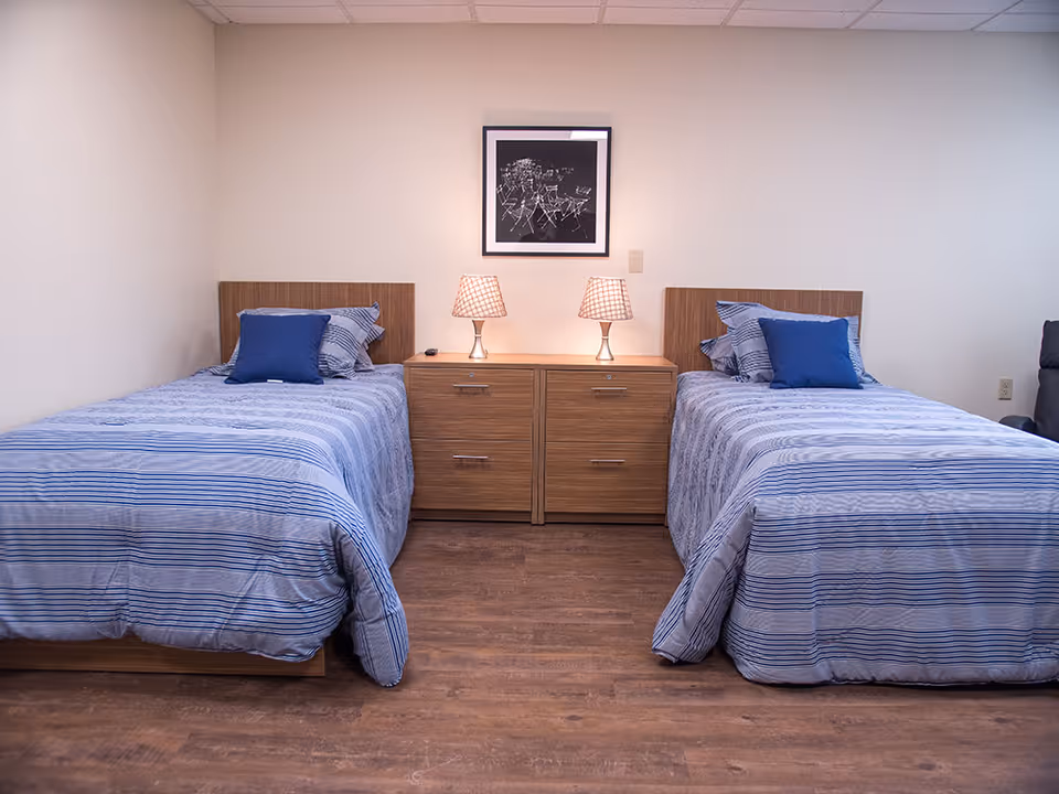 A bedroom with two twin beds separated by two wooden nightstands with drawers. Each bed has blue and white striped bedding and a blue pillow. Two table lamps with checkered shades are on the nightstands. A framed black and white photograph hangs on the wall above the nightstands. The floor is wooden and the walls are plain white.