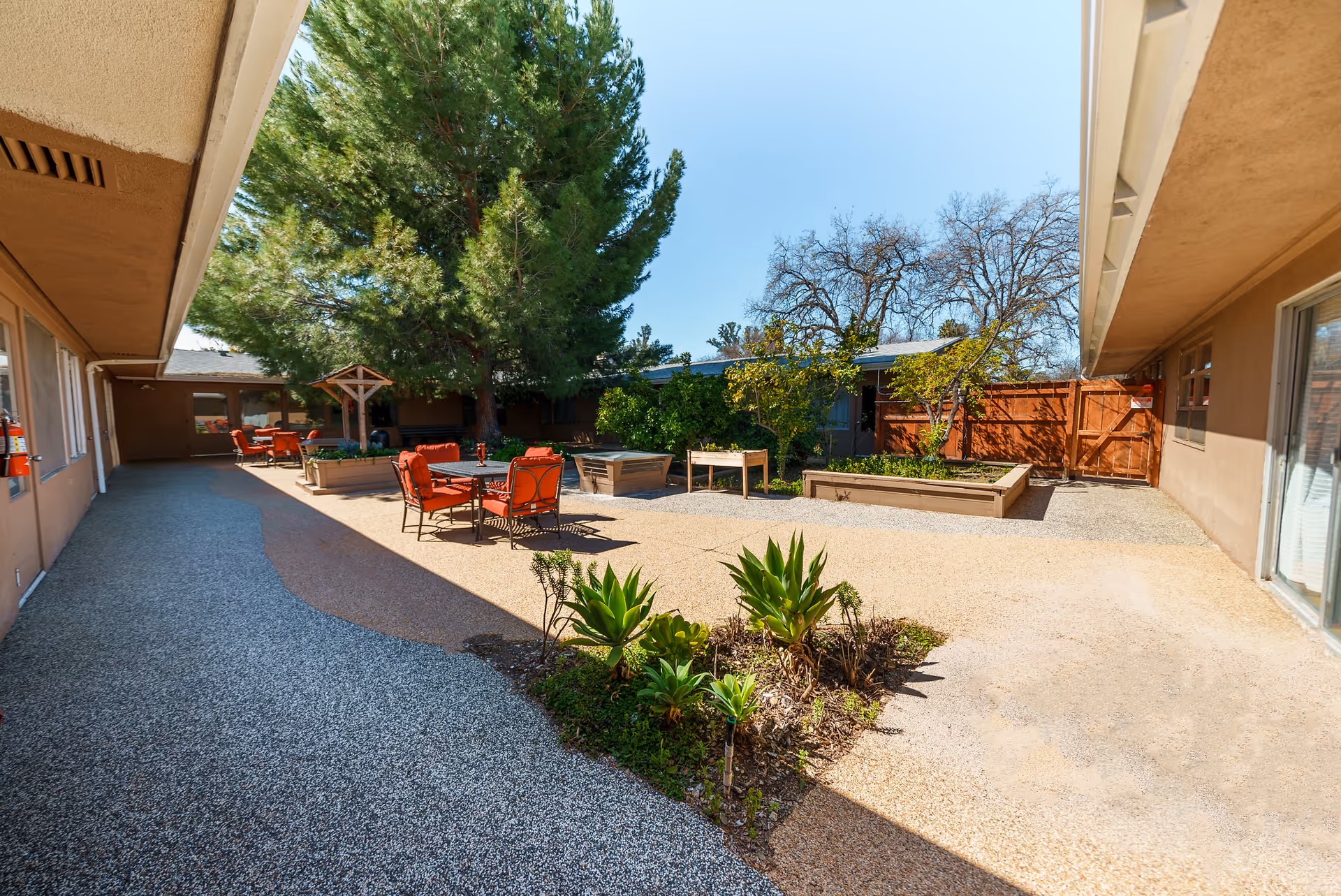 Outdoor courtyard area at Ojai Health & Rehabilitation featuring several red cushioned chairs and tables, raised garden beds with plants, a large tree providing shade, and a clear blue sky overhead.