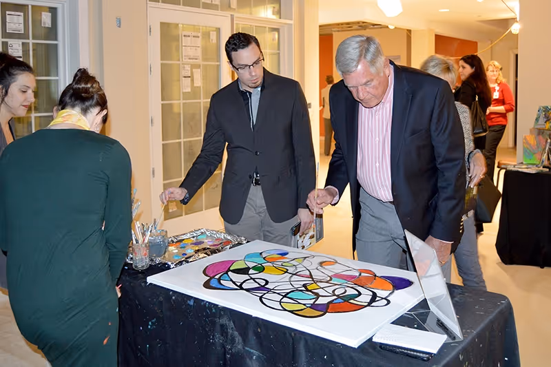 A group of people gathered around a table displaying a colorful abstract painting. One man in a suit is closely examining the artwork while others look on. The setting appears to be an indoor event or gallery space with bright lighting and a few other artworks visible in the background.