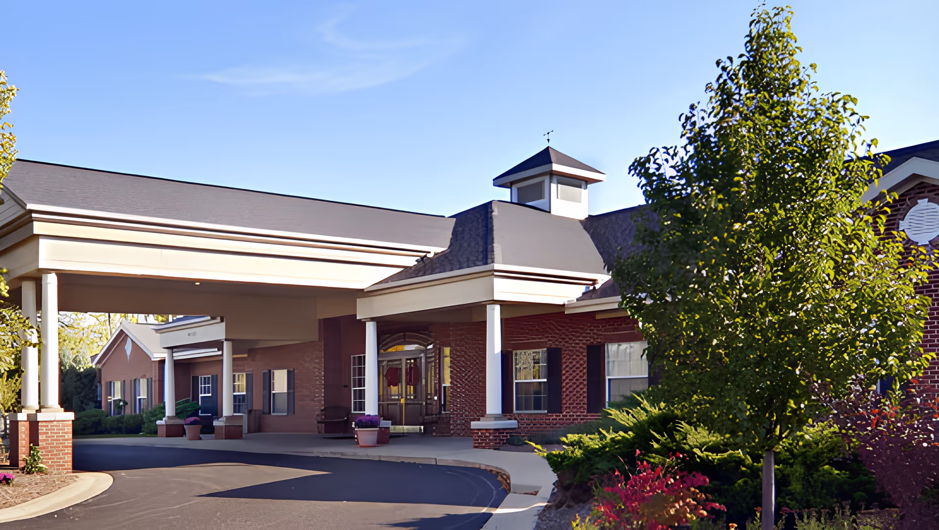 Exterior view of Care Cardinal Kentwood facility showing a covered entrance with white columns, brick walls, and a landscaped area with trees and shrubs under a clear blue sky.