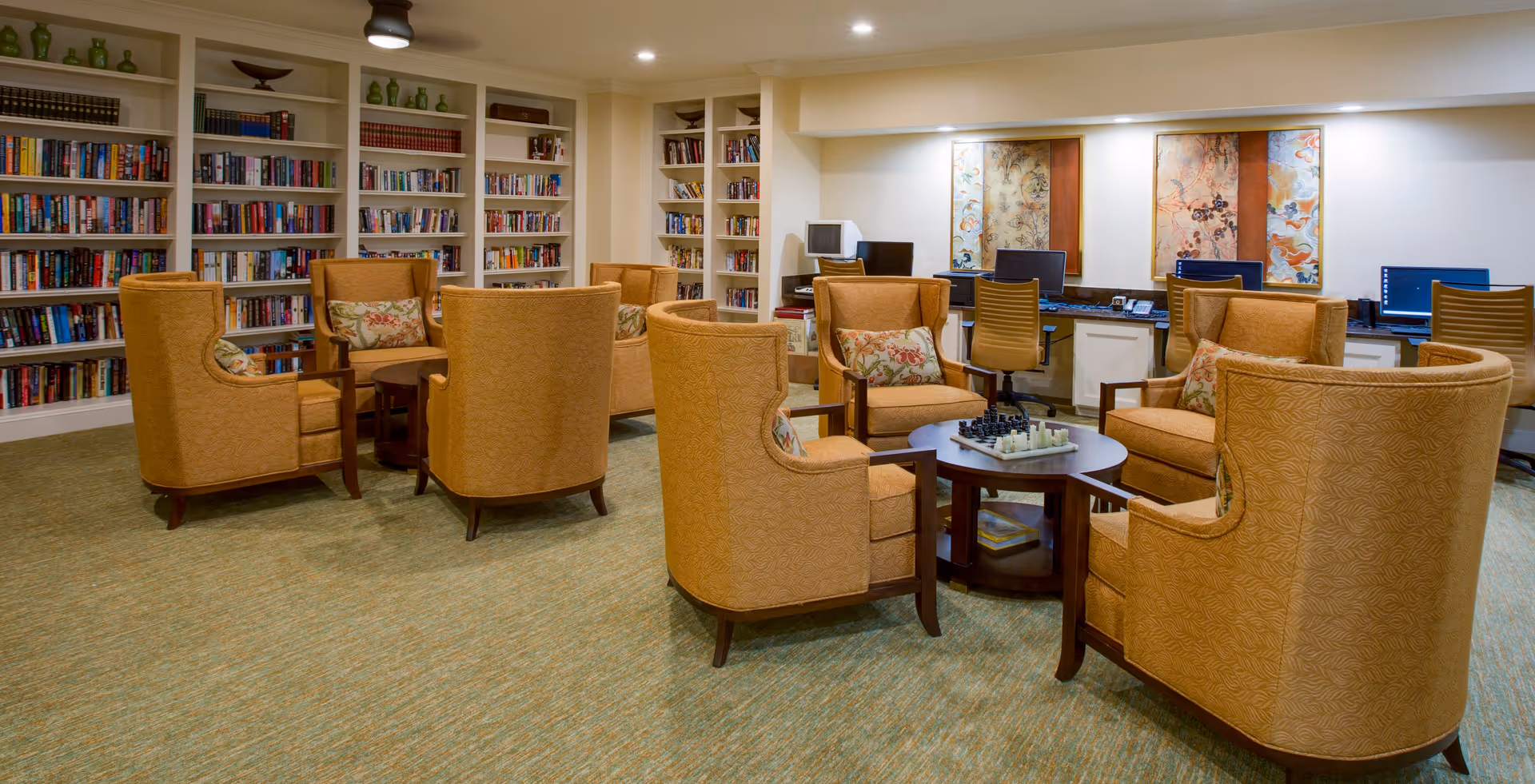A cozy library and computer area in a senior living facility featuring multiple bookshelves filled with books, several comfortable upholstered armchairs arranged around small tables with a chess set, and a row of desks with computers and office chairs against the wall. The room is well-lit with recessed ceiling lights and decorated with framed artwork.