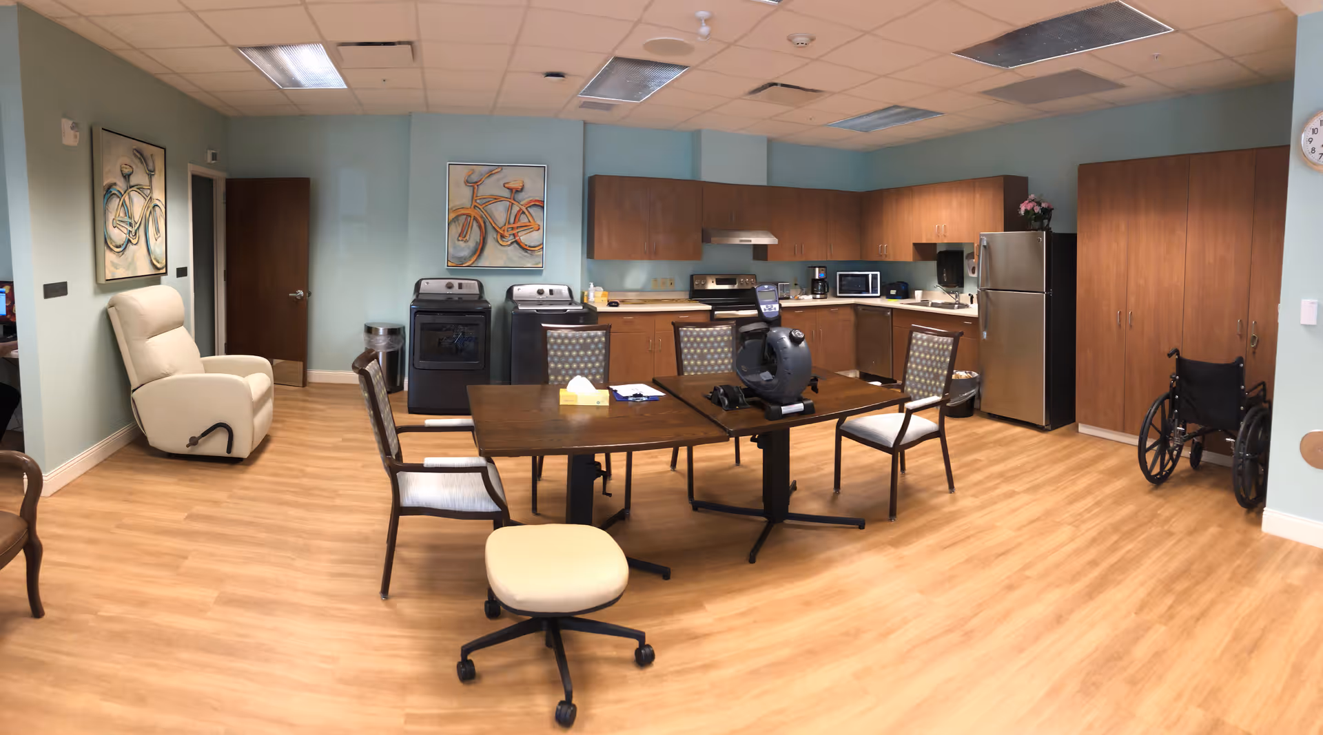 A spacious room with light wood flooring and light blue walls featuring a kitchen area with wooden cabinets, a refrigerator, microwave, stove, and dishwasher. In the center, there is a wooden table with four chairs and a small rolling stool. A beige recliner chair is positioned near a closed wooden door on the left side. A wheelchair is placed near a large wooden cabinet on the right side. The walls are decorated with two paintings of bicycles. The ceiling has recessed lighting and ventilation panels.