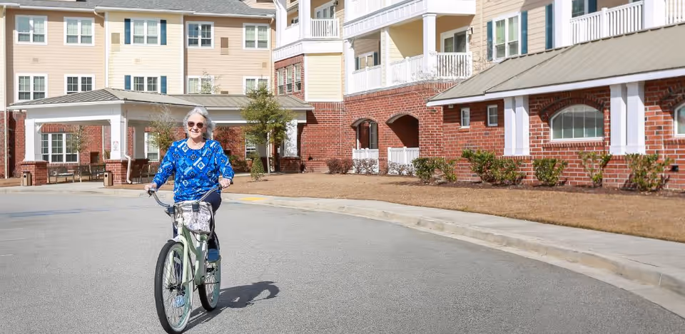 A person rides a bicycle on the driveway in front of a multi-story senior living building with brick lower walls and light-colored siding.