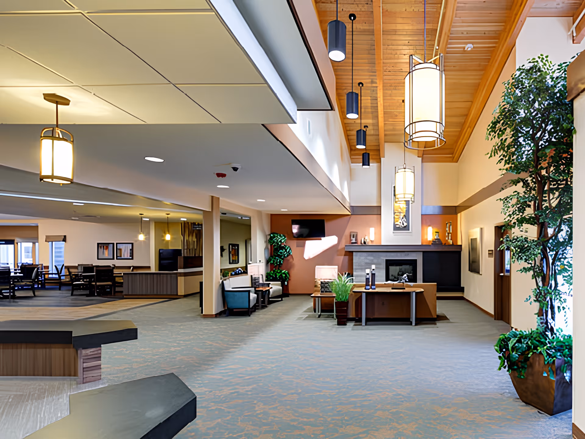 Spacious and well-lit interior common area of a senior living facility featuring a high wooden ceiling with hanging pendant lights, a fireplace with a mantle, seating areas with chairs and tables, potted plants, and a dining area visible in the background.