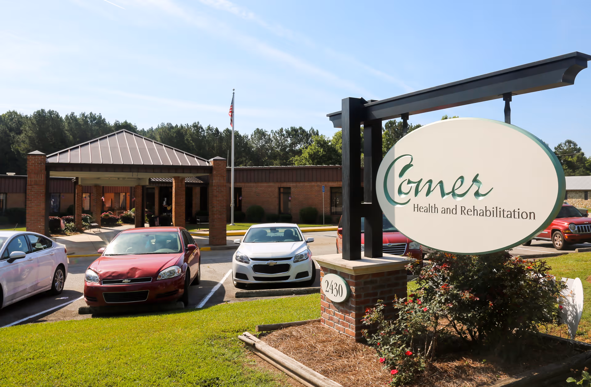 Exterior view of Comer Health and Rehabilitation facility with a large sign displaying the facility name in the foreground. Several cars are parked in front of the single-story brick building with a covered entrance and an American flag on a flagpole.
