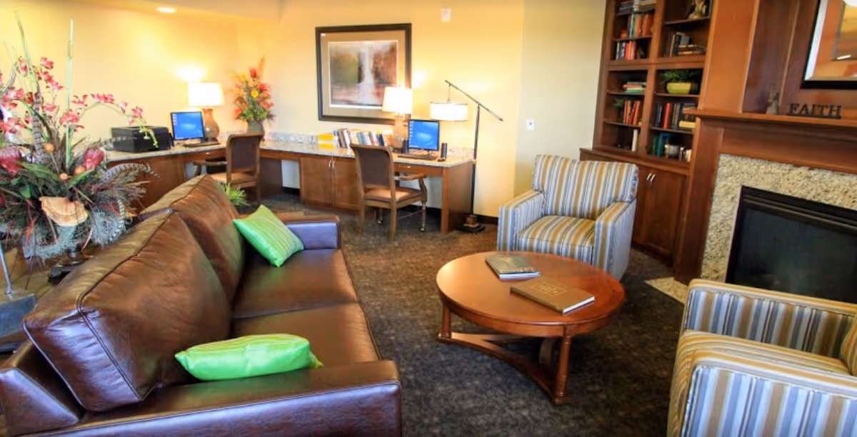 A cozy living room area with a brown leather sofa adorned with green pillows, two striped armchairs, and a round wooden coffee table with books on it. In the background, there is a wooden desk with two chairs, computers, lamps, and a framed picture on the wall. To the right, there is a fireplace with built-in wooden shelves filled with books and decorative items.