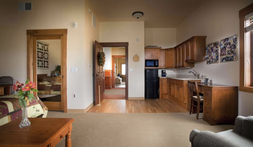 Interior view of a senior living facility room featuring a small kitchenette with wooden cabinets, a black refrigerator, microwave, and coffee maker. To the left, there is a bed with a floral quilt partially visible, a wooden table with a vase of flowers, and a doorway leading to another room. On the right, there is a wooden desk with a chair beneath a window, and a comfortable armchair in the foreground. The walls are light-colored, and the floor is a combination of carpet and wood.