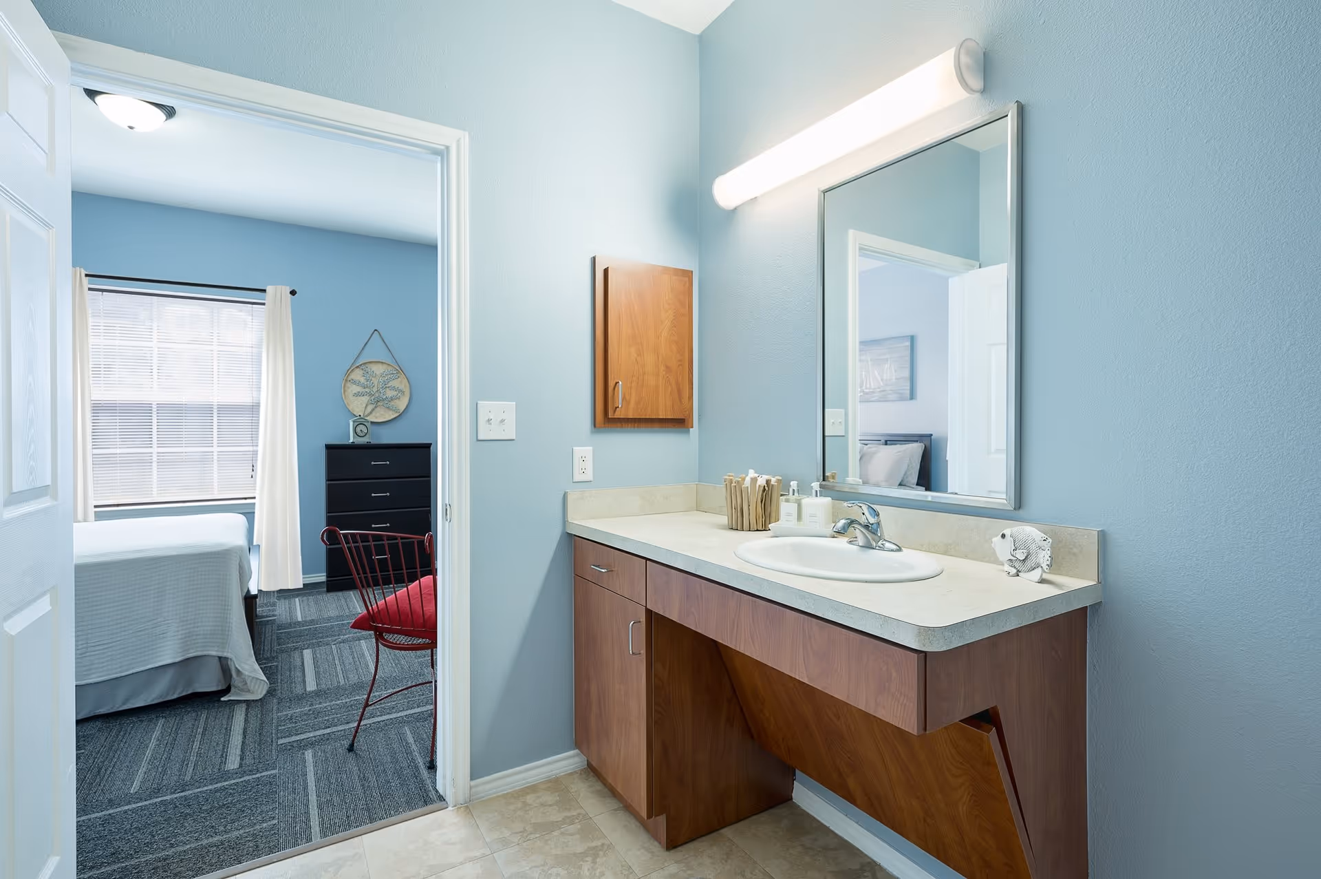 A bathroom vanity area with a large mirror, a sink, and wooden cabinets. The walls are painted light blue, and there is a light fixture above the mirror. Through an open door, a bedroom with a bed, a window with white curtains, a black dresser, and a red chair is visible.