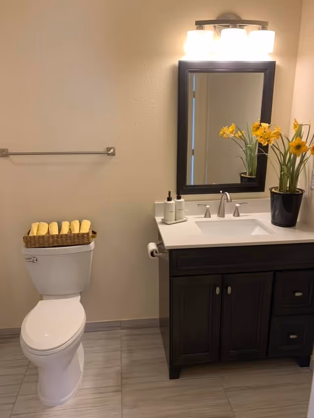 A clean bathroom with a white toilet topped with a basket holding rolled yellow towels. Next to the toilet is a dark wood vanity with a white countertop, a silver faucet, two soap dispensers, and a black pot with yellow flowers. Above the vanity is a rectangular mirror with a black frame and a light fixture with three bulbs.
