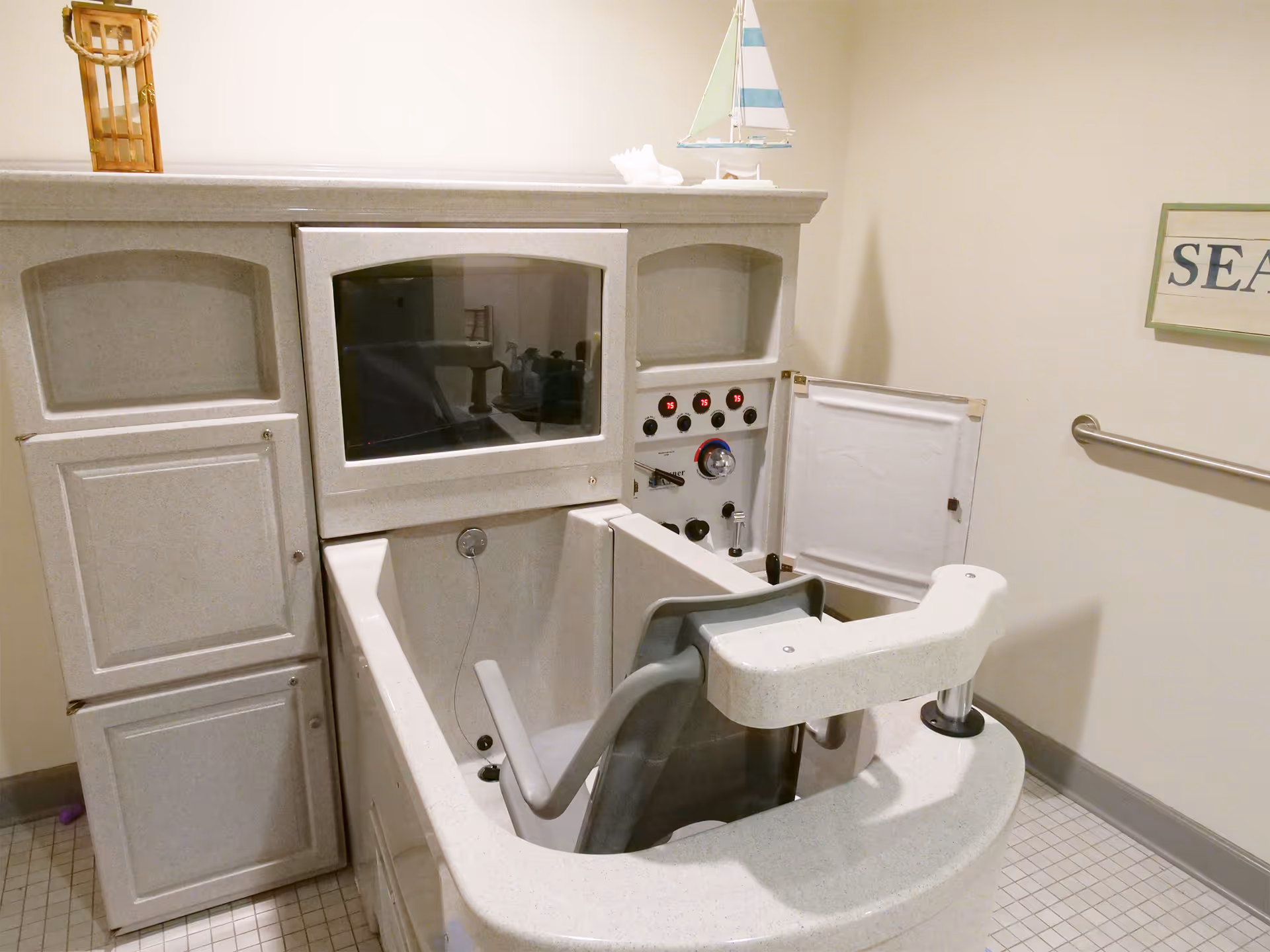 A specialized assisted bathing tub with a built-in chair and control panel, located in a tiled room with a grab bar on the wall and a decorative sailboat and lantern on top of the tub enclosure.