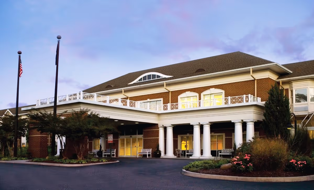 Exterior front view of Shalom Gardens Senior Assisted Living Facility in Virginia during dusk, showing a large brick building with white columns, a covered entrance, landscaped garden beds with flowers and shrubs, and two flagpoles with flags.